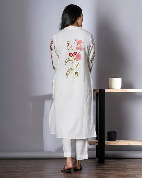 Woman wearing a white embroidered outfit standing in a minimalistic room.