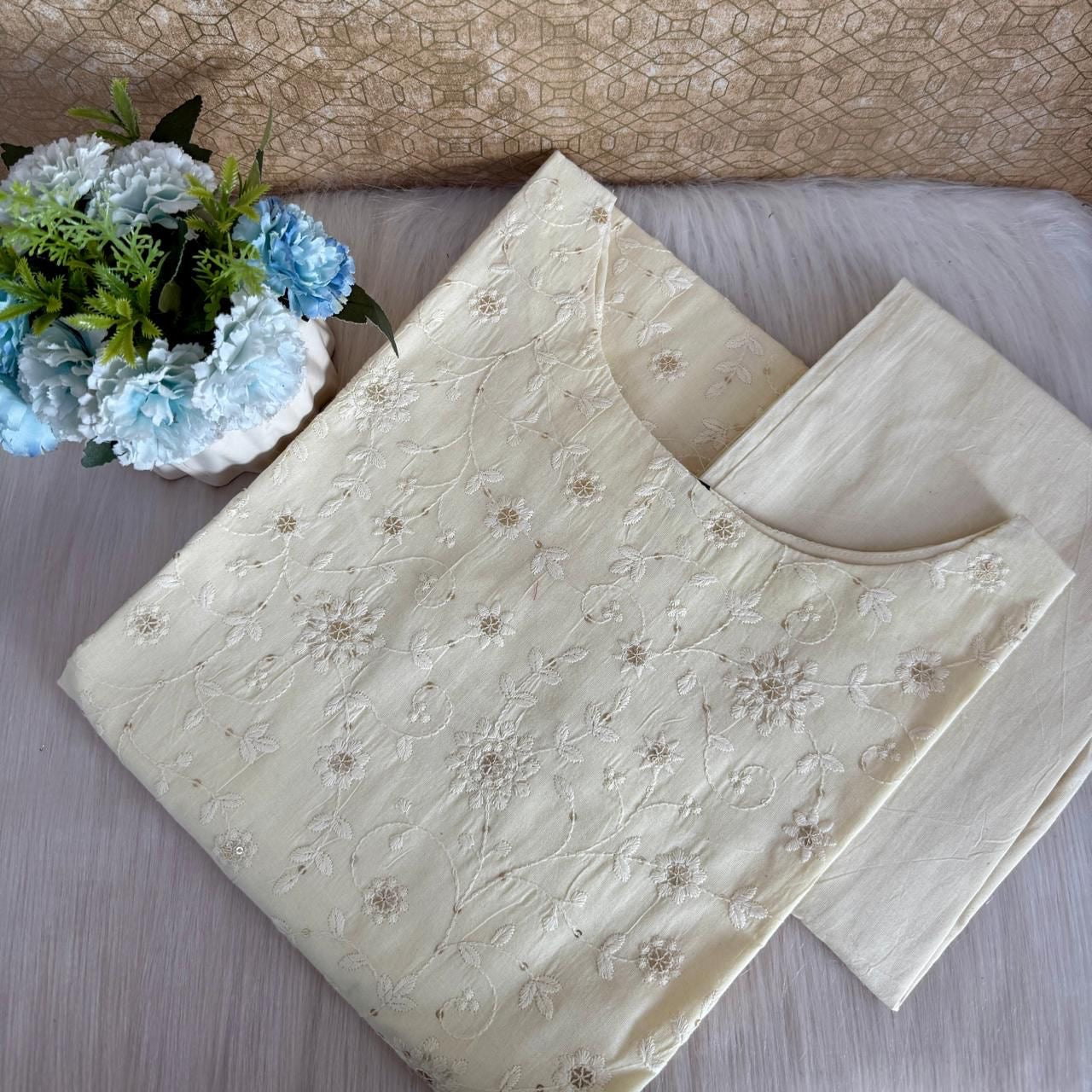 Beige embroidered fabric with a floral pattern on a wooden surface, next to a small potted plant with blue flowers.