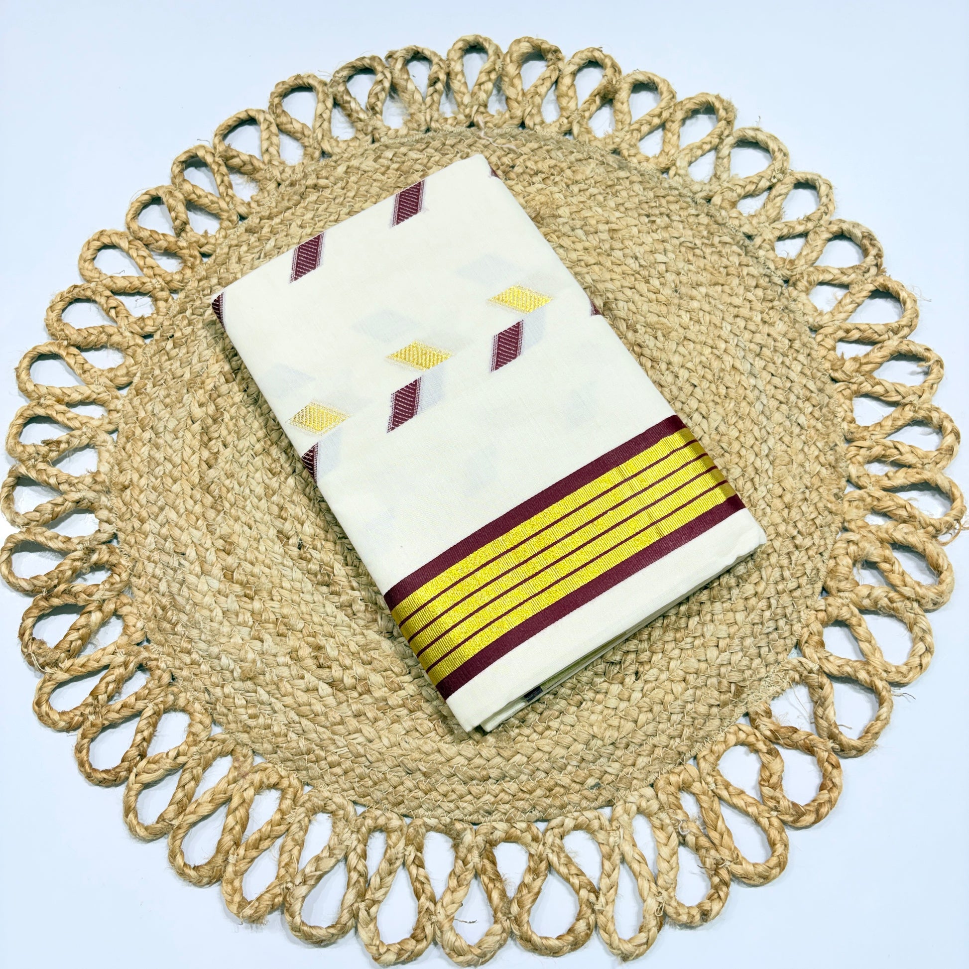 Off-white Kerala cotton saree with multicolour diamond butta motifs and yellow-maroon stripe border, neatly folded on a jute woven placemat – perfect for Onam, festivals and office wear