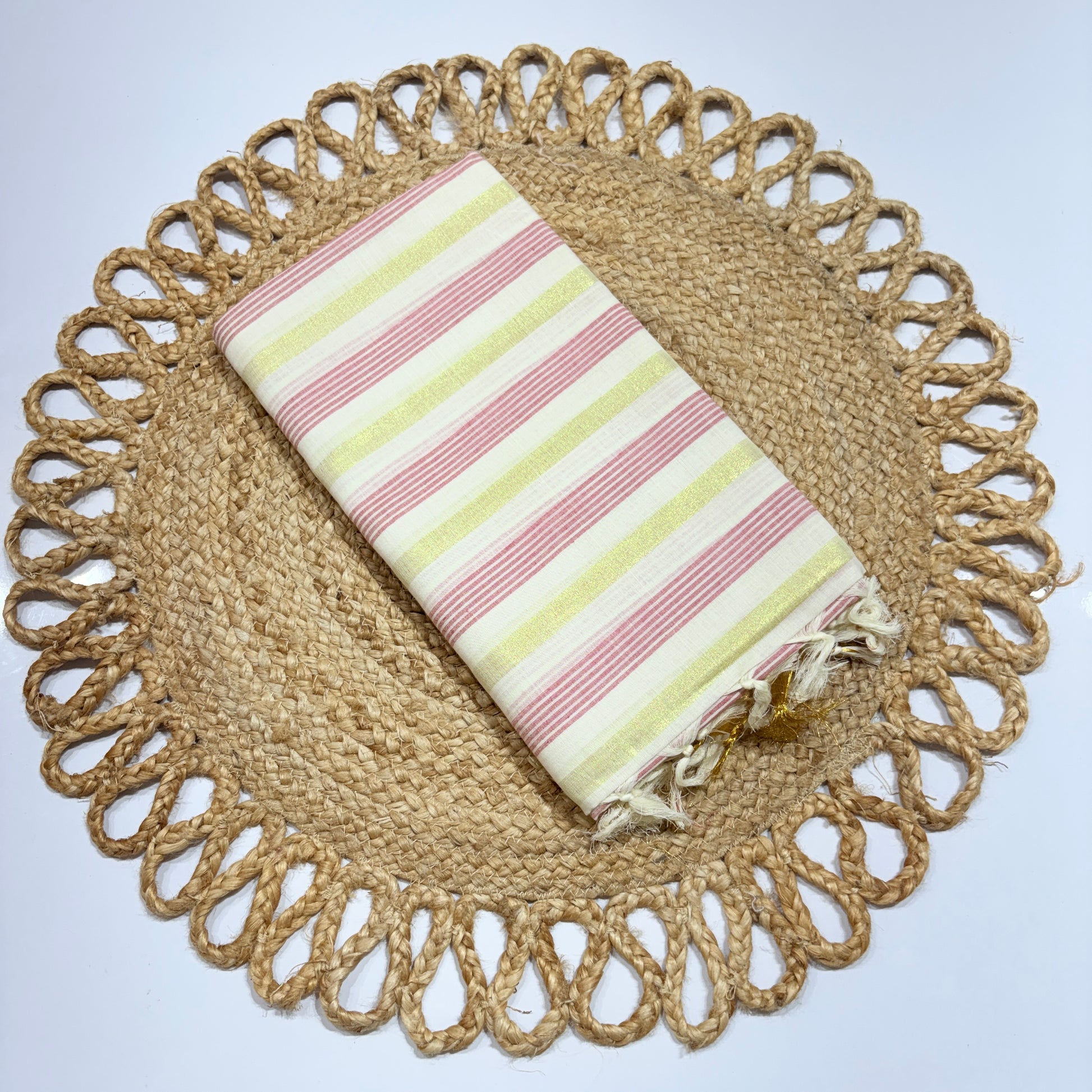 Kerala Cotton Saree Off-White with Pink & Yellow 6-Line Zari Stripe Weave and Gold Tassel Pallu – neatly folded on a jute woven placemat, lightweight Onam and festival saree by RAE Trends