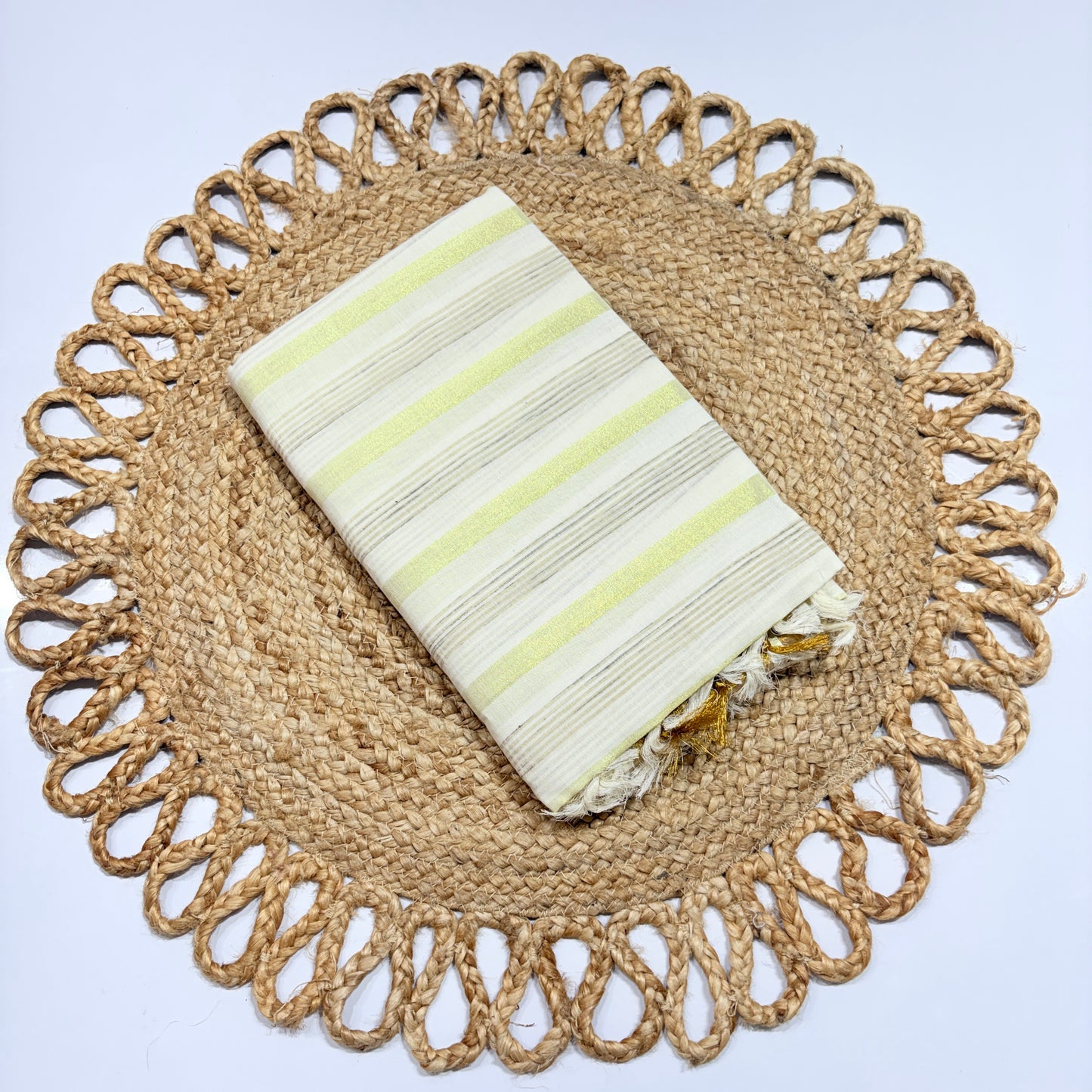 Kerala Cotton Saree Off-White with Yellow Zari & Silver Stripe Weave and Gold Tassel Pallu – neatly folded on a jute woven placemat, lightweight Onam and festival saree by RAE Trends