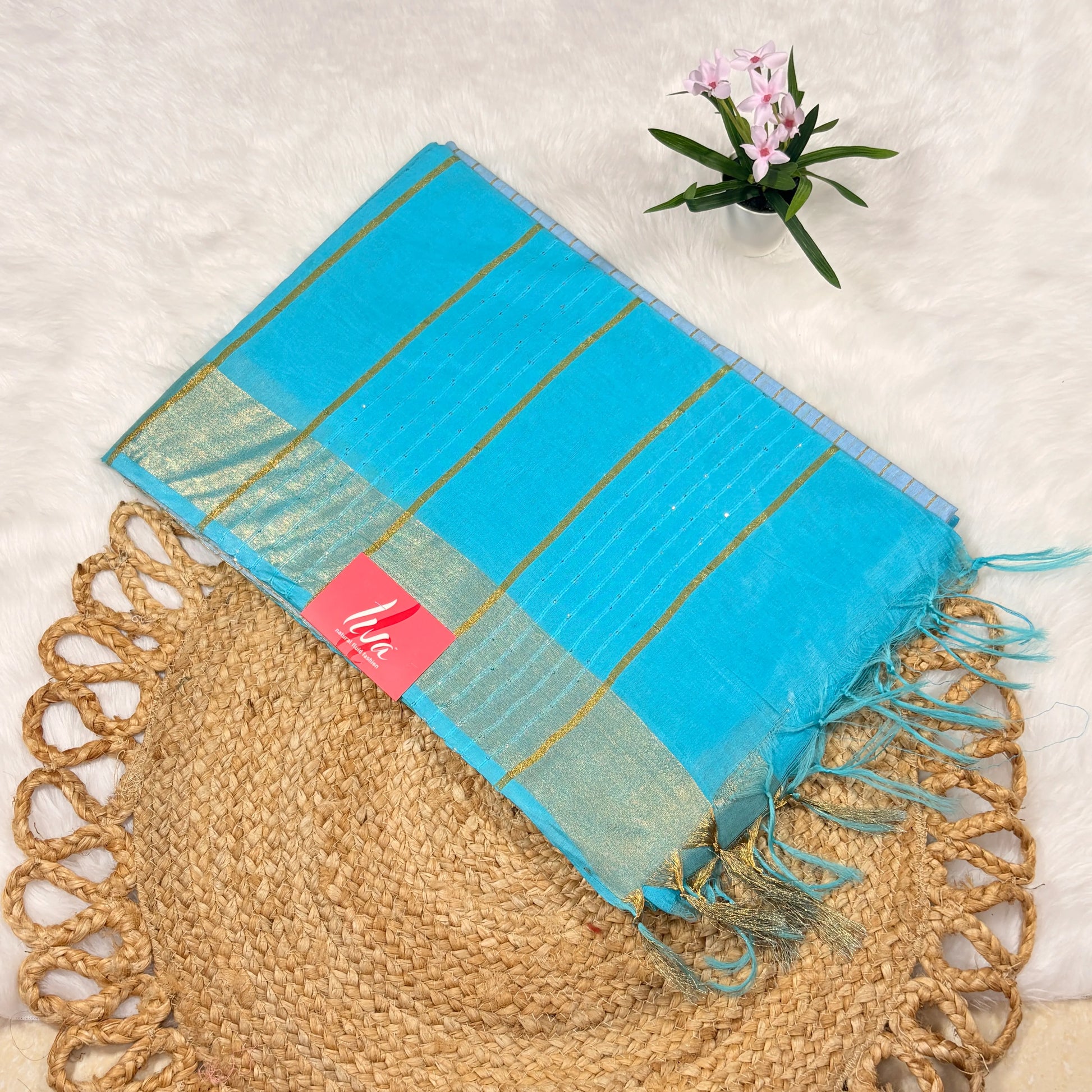Turquoise scarf with gold stripes and tassels on a textured surface with a plant in the corner.