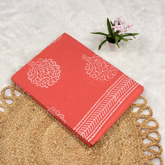 Coral cotton Jaipur block print saree with white paisley scroll motifs, leaf‑chevron border lines, and matching blouse piece folded on a woven mat