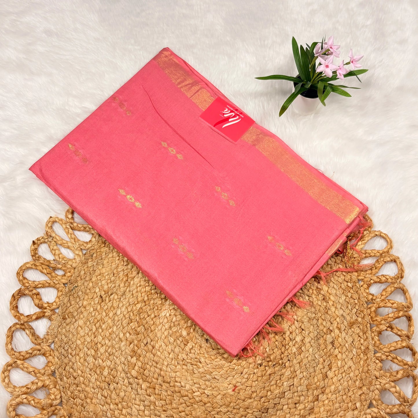 Rose‑pink Liva saree with scattered zari buti motifs, linear jari pallu, tasselled edge, and matching blouse piece, folded on a natural mat.