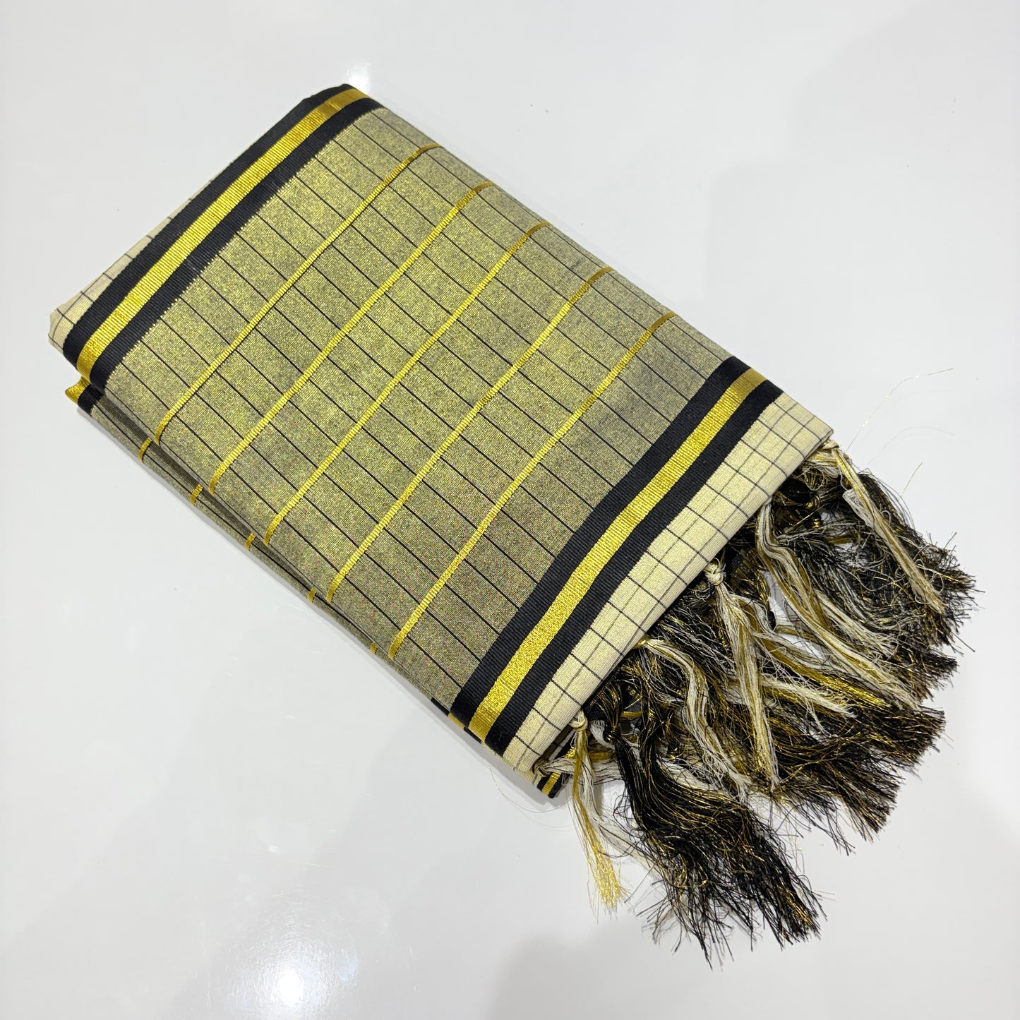 Kerala tissue silk saree neatly folded showing gold and black check weave body with yellow zari accents, bold black and gold stripe border, and dramatic black and gold mixed tassel fringe pallu – premium festive saree