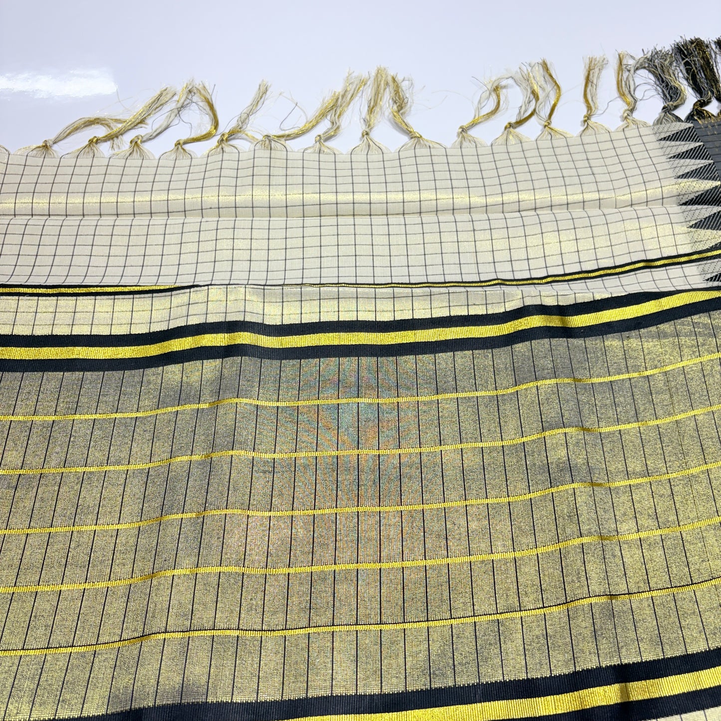 Close-up of Kerala tissue silk saree showing gold shimmer check weave body with fine black grid lines, yellow zari horizontal stripes on pallu, bold black and gold stripe border, and cream check body with gold tassel fringe – Onam and wedding wear